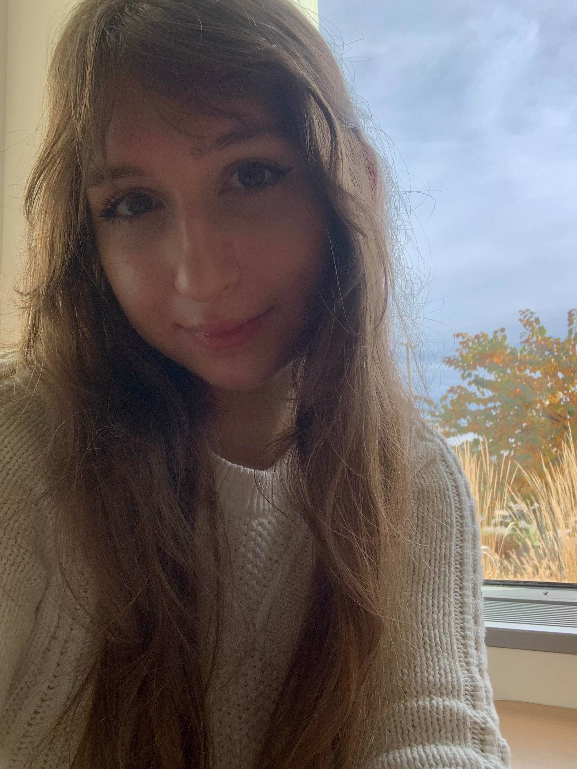 Girl with long, brown hair smiling at the camera with blue sky and trees in the background